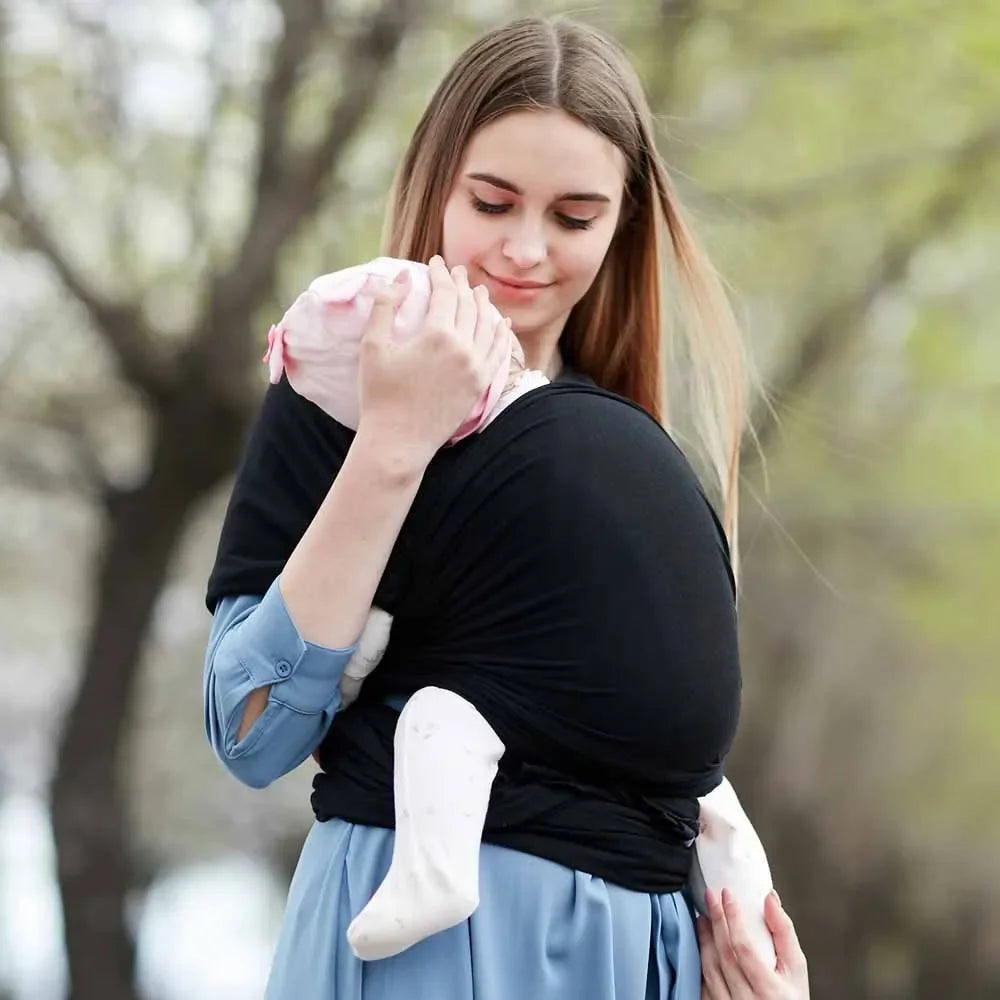 A baby sling wrap carrier with a sleeping infant inside, being worn by an adult with one hand supporting the baby's head.